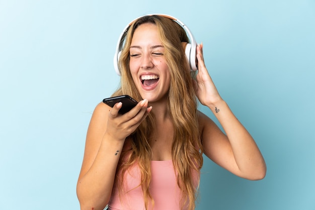 Giovane donna bionda isolata sulla musica d'ascolto della parete blu con un cellulare e cantando