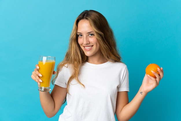 Giovane donna bionda isolata sulla parete blu che tiene un'arancia e un succo d'arancia