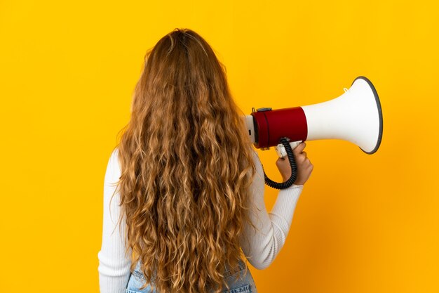 Giovane donna bionda isolata sulla parete gialla che tiene un megafono e nella posizione posteriore