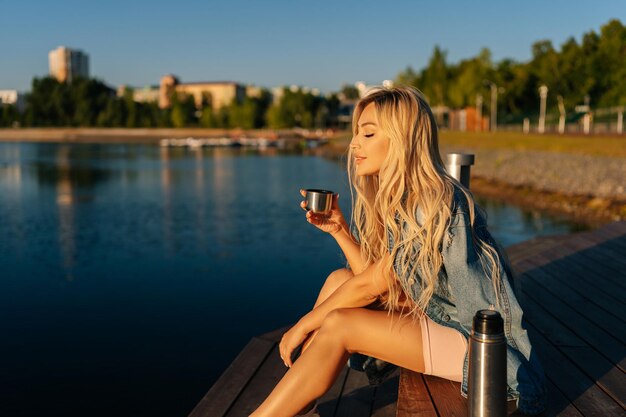 Giovane donna bionda rilassata che tiene una tazza con caffè caldo dal thermos seduto sulla spiaggia vicino al molo di legno la mattina di sole estivo
