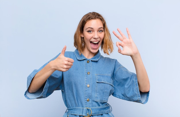 Giovane donna bionda sentirsi felice, stupito, soddisfatto e sorpreso, mostrando ok e pollice in alto gesti, sorridendo contro la parete di colore piatto