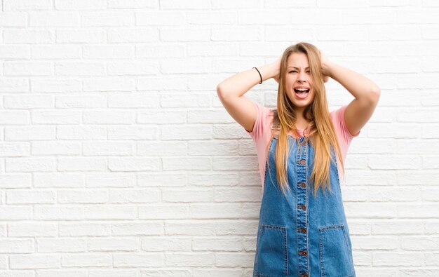 Giovane donna bionda sentirsi stressata, preoccupata, ansiosa o spaventata, con le mani sulla testa, in preda al panico per errore contro il muro di mattoni