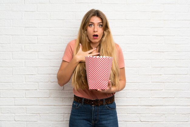 Giovane donna bionda sopra il muro di mattoni bianchi che tiene una ciotola di popcorn