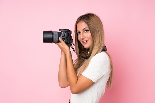 Giovane donna bionda sopra il rosa isolato con una macchina fotografica professionale