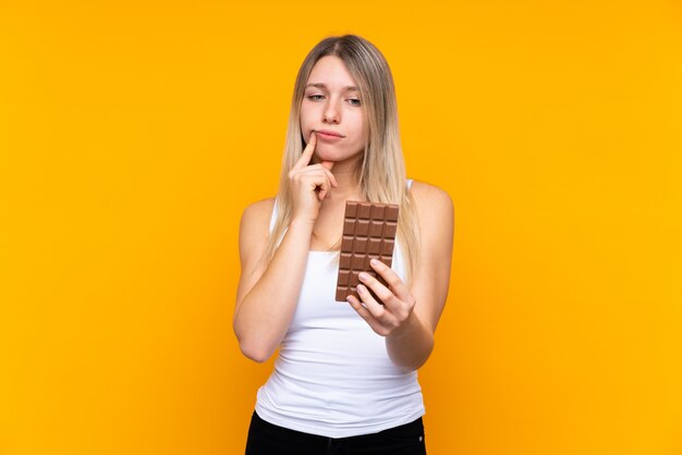 Giovane donna bionda sopra la parete blu isolata che prende una compressa del cioccolato e che ha dubbi