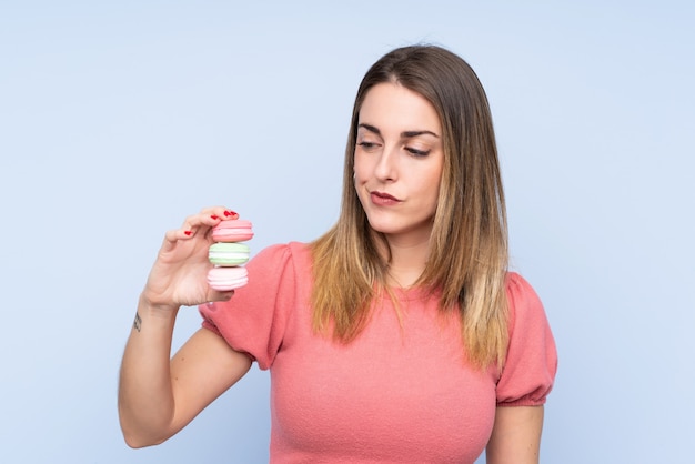 Giovane donna bionda sopra la parete blu isolata che tiene i macarons francesi variopinti con l'espressione triste