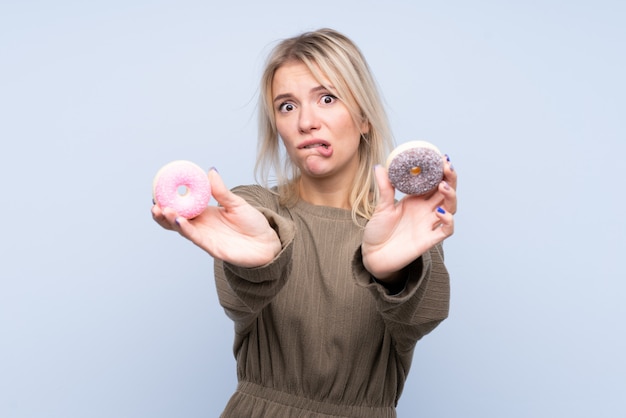 Giovane donna bionda sopra la parete blu isolata che tiene una ciambella e triste