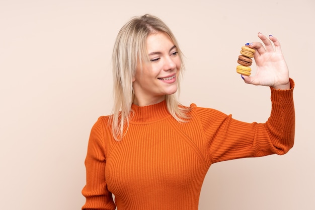 Giovane donna bionda sopra la parete isolata che tiene macarons francesi variopinti e con l'espressione felice