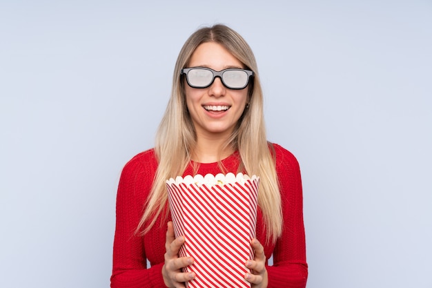 Giovane donna bionda sopra la parete isolata con gli occhiali 3d e in possesso di un grande secchio di popcorn