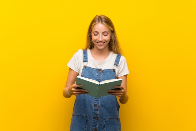 Giovane donna bionda sopra la tenuta gialla isolata della parete e la lettura del libro