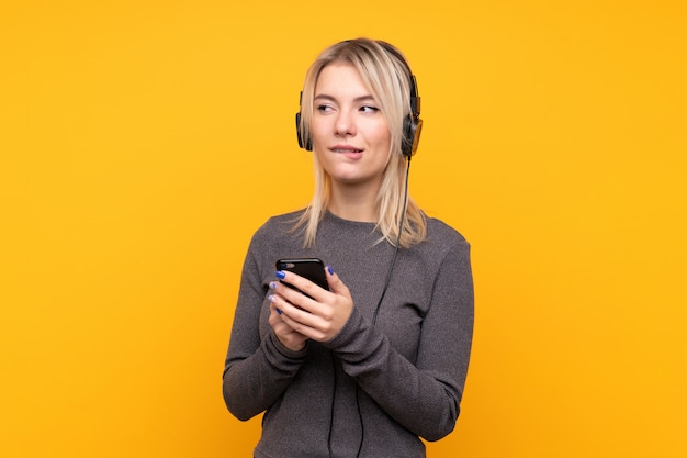 Giovane donna bionda sopra musica d'ascolto isolata della parete gialla con un cellulare e un pensiero