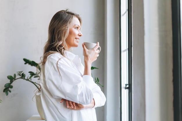 Giovane donna bionda sorridente con i capelli lunghi in camicia bianca riposa e beve il tè vicino alla finestra nell'ufficio moderno e luminoso