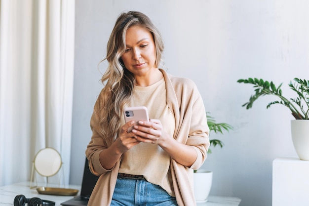 Giovane donna bionda sorridente con i capelli lunghi in cardigan elegante che lavora al computer portatile nel luminoso ufficio moderno donna invia un messaggio tramite smartphone