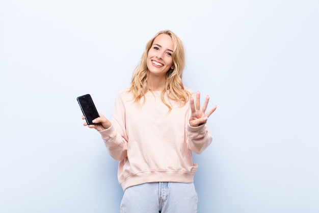 Giovane donna bionda sorridente e dall'aspetto amichevole, mostrando il numero quattro o quarto con la mano in avanti, il conto alla rovescia utilizzando uno smartphone