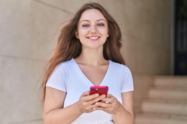 Giovane donna bionda sorridente fiducioso utilizzando lo smartphone in strada