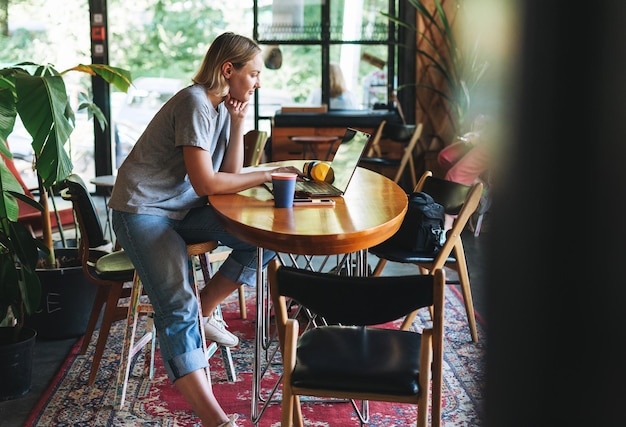 Giovane donna bionda sorridente freelance con le cuffie gialle che lavora al portatile sul tavolo del caffè
