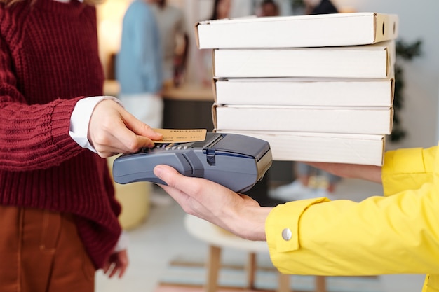 Giovane donna bionda sorridente in abbigliamento casual intelligente in possesso di carta di credito sullo schermo della macchina di pagamento in mano del corriere maschio in uniforme gialla