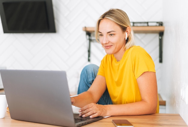 Giovane donna bionda sorridente in maglietta gialla con le cuffie che lavorano al computer portatile in cucina a casa