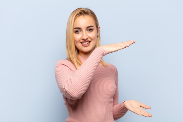 Giovane donna bionda sorridente, sentendosi felice, positivo e soddisfatto, tenendo o mostrando un oggetto o un concetto