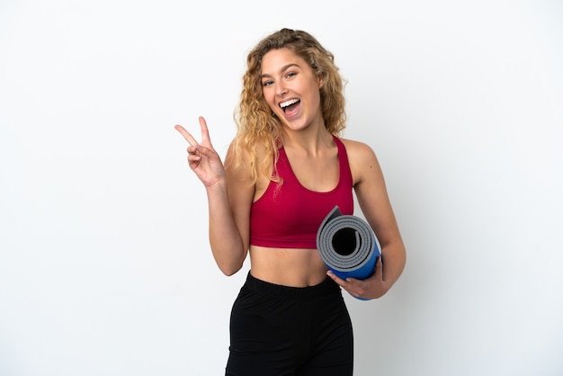 Giovane donna bionda sportiva che va alle lezioni di yoga mentre tiene in mano un tappetino isolato su sfondo bianco sorridendo e mostrando il segno della vittoria