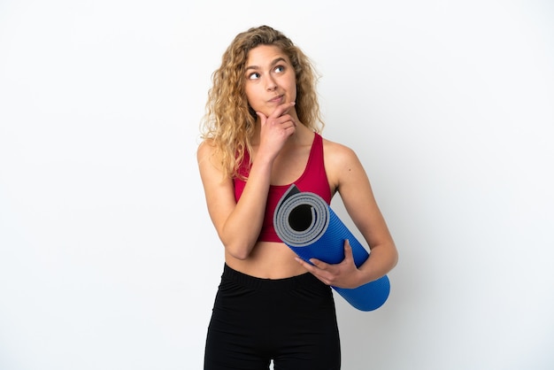 Giovane donna bionda sportiva che va alle lezioni di yoga tenendo in mano un tappetino isolato su sfondo bianco e guardando in alto