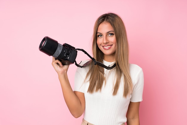 Giovane donna bionda su sfondo rosa isolato con una macchina fotografica professionale