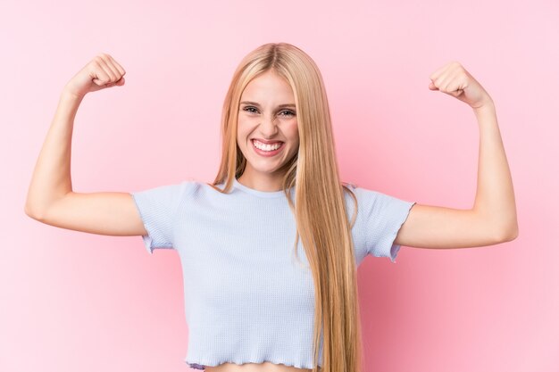 Giovane donna bionda sulla parete rosa che mostra il gesto di forza con le braccia, simbolo del potere femminile