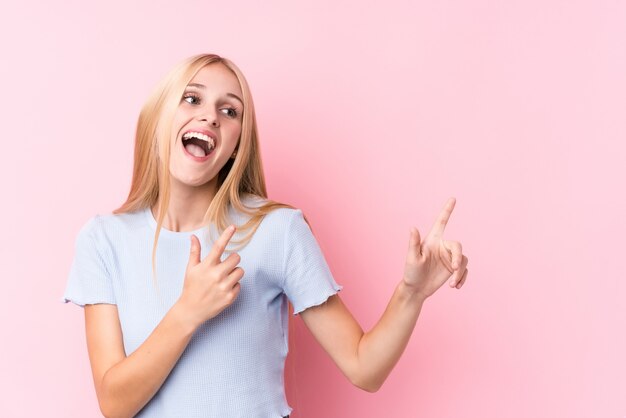 Giovane donna bionda sulla parete rosa che punta con gli indici uno spazio di copia, esprimendo eccitazione e desiderio.