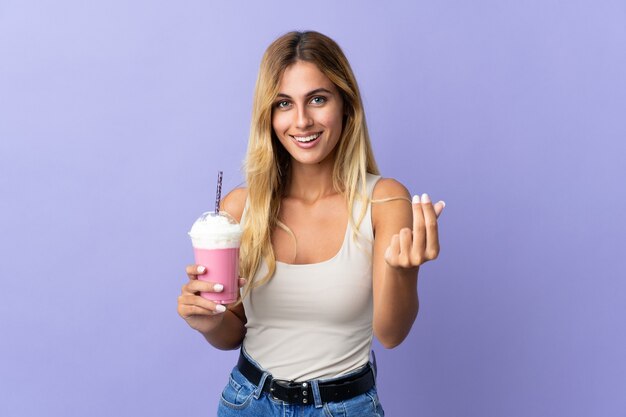 Giovane donna bionda uruguaiana con frappè alla fragola sul gesto di fare soldi viola