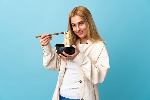 Giovane donna bionda uruguaiana sopra l'azzurro isolato che tiene una ciotola di tagliatelle con le bacchette