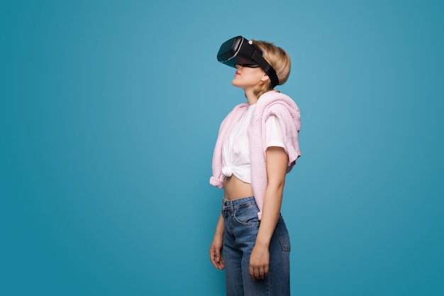 Giovane donna bionda utilizzando le cuffie da realtà virtuale e in posa su una parete blu