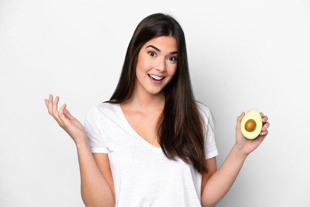 Giovane donna brasiliana che tiene un avocado isolato su sfondo bianco con espressione facciale scioccata