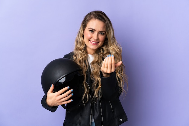 Giovane donna brasiliana che tiene un casco del motociclo isolato su sfondo viola che invita a venire con la mano. Felice che tu sia venuto