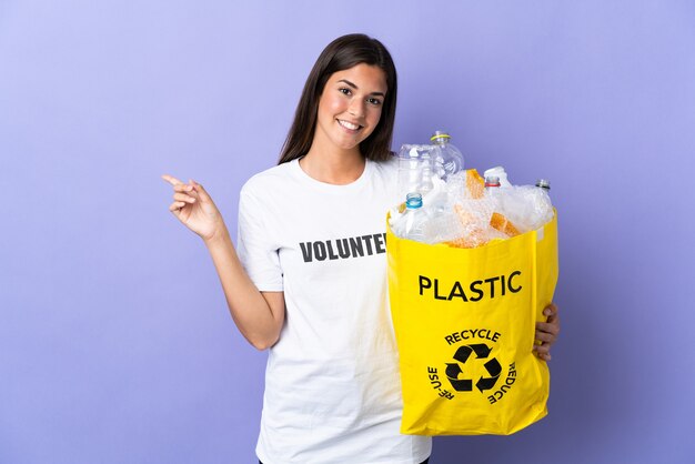 Giovane donna brasiliana che tiene un sacchetto pieno di bottiglie di plastica da riciclare isolato sul dito puntato viola a lato