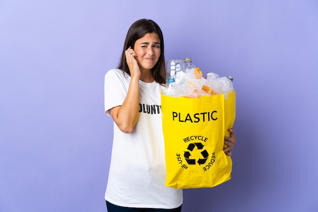Giovane donna brasiliana che tiene un sacchetto pieno di bottiglie di plastica da riciclare isolato sulle orecchie frustrate e coniche viola