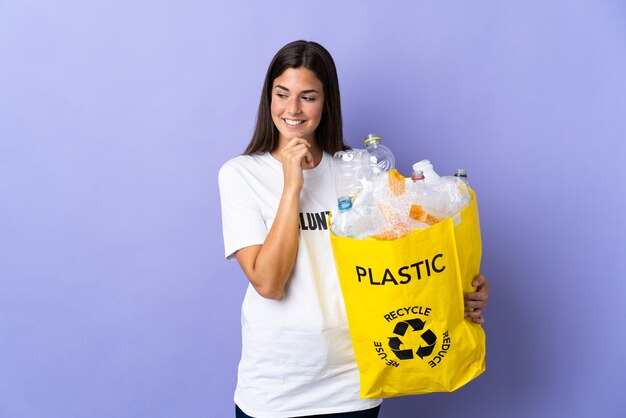 Giovane donna brasiliana che tiene una borsa piena di bottiglie di plastica da riciclare isolata sulla parete viola che guarda al lato e che sorride