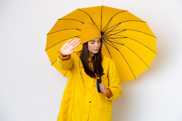 Giovane donna brasiliana con cappotto antipioggia e ombrello isolati su sfondo bianco che fa un gesto di arresto e delusa
