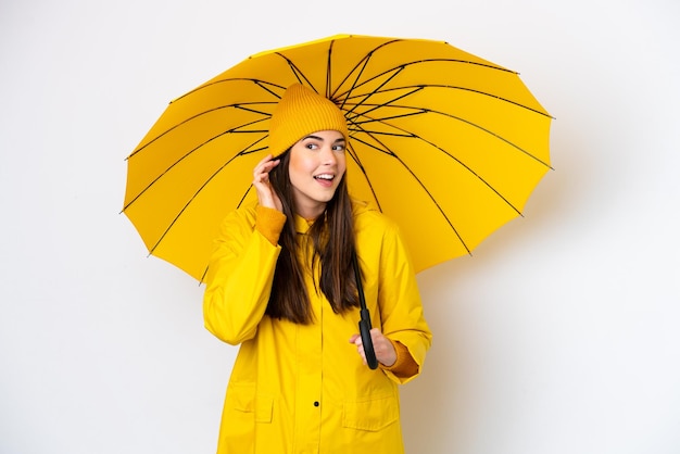 Giovane donna brasiliana con cappotto antipioggia e ombrello isolato su sfondo bianco ascoltando qualcosa mettendo la mano sull'orecchio