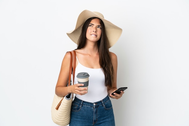 Giovane donna brasiliana con Pamela che tiene in mano una borsa da spiaggia isolata su sfondo bianco che tiene il caffè da portare via e un cellulare mentre pensa a qualcosa