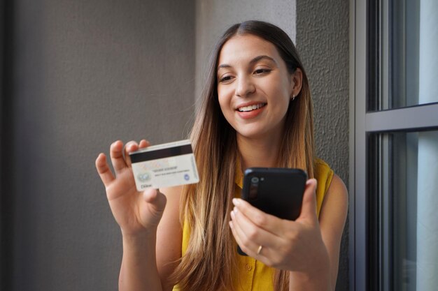 Giovane donna brasiliana felice con uno smartphone e che legge il numero della carta di credito per fare acquisti online