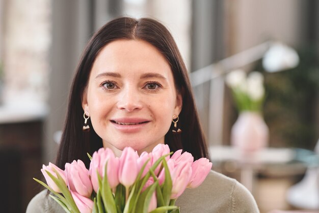 Giovane donna bruna allegra che ti guarda con un sorriso mentre tiene un mazzo di tulipani rosa freschi per il mento e si gode la giornata di primavera