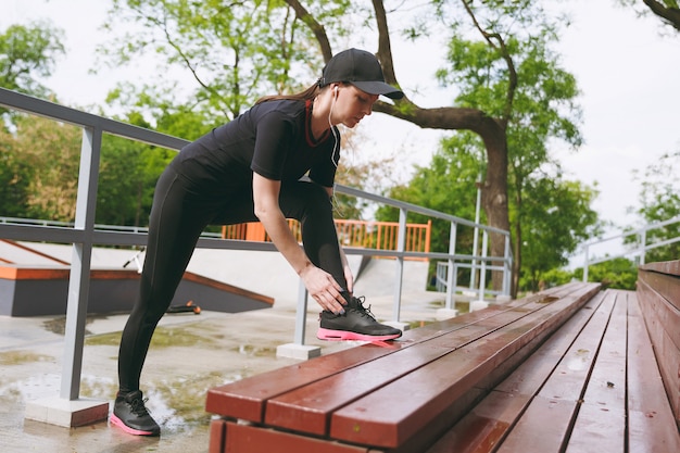 Giovane donna bruna atletica concentrata in uniforme nera, berretto con auricolari che ascolta musica facendo esercizi di stretching sportivo riscaldamento sulla panchina nel parco cittadino all'aperto