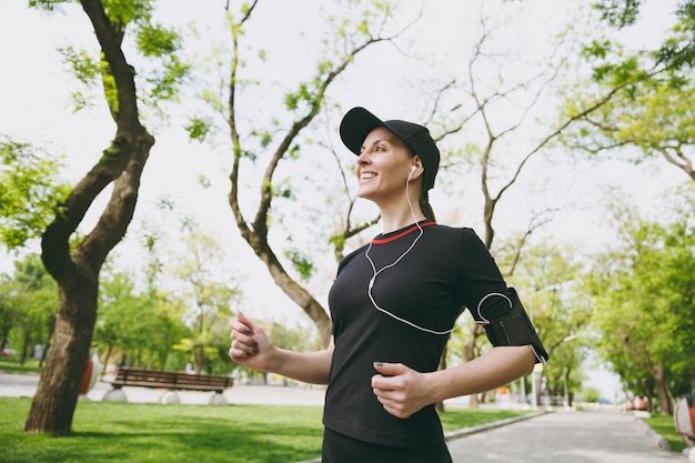 Giovane donna bruna atletica sorridente in uniforme nera e berretto con auricolari che si allenano facendo sport, correndo e ascoltando musica sul sentiero nel parco cittadino all'aperto