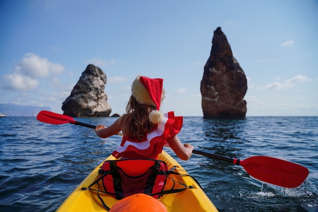 Giovane donna bruna attraente in costume da bagno rosso e cappello da Babbo Natale che nuota in kayak intorno a rocce di basalto