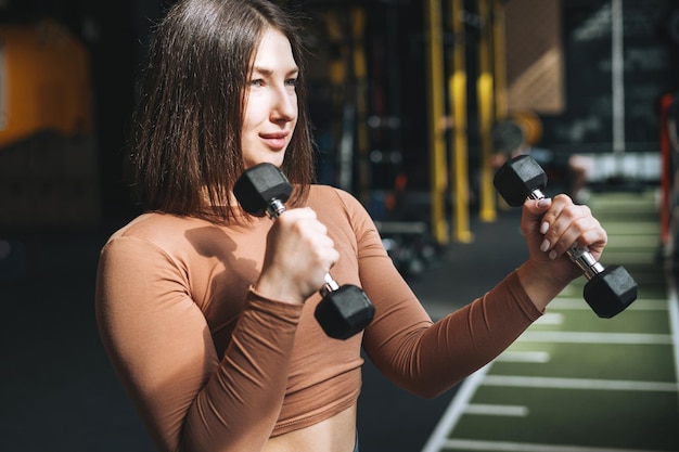 Giovane donna bruna che allena i suoi muscoli con i manubri nella palestra del fitness club