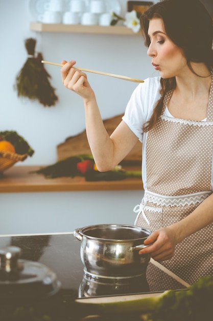 Giovane donna bruna che cucina minestra in cucina. Casalinga che tiene in mano un cucchiaio di legno. Concetto di cibo e salute.