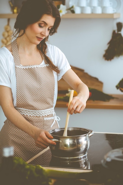 Giovane donna bruna che cucina minestra in cucina. Casalinga che tiene in mano un cucchiaio di legno. Concetto di cibo e salute.