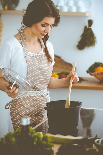 Giovane donna bruna che cucina minestra in cucina. Casalinga che tiene in mano un cucchiaio di legno. Concetto di cibo e salute.