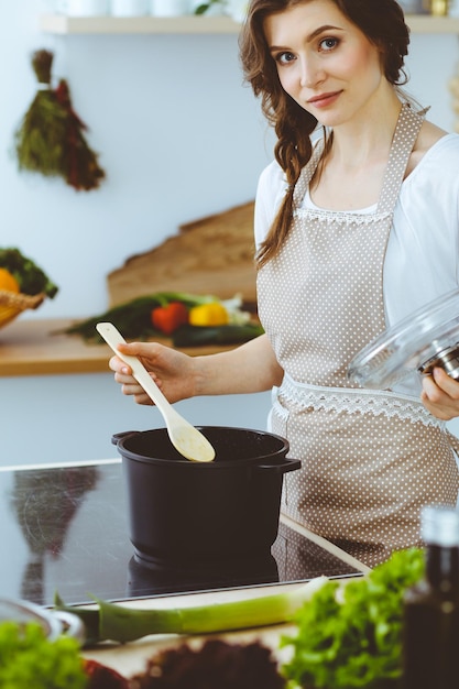 Giovane donna bruna che cucina minestra in cucina. Casalinga che tiene in mano un cucchiaio di legno. Concetto di cibo e salute.