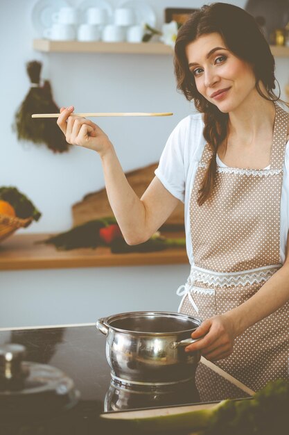 Giovane donna bruna che cucina minestra in cucina. Casalinga che tiene in mano un cucchiaio di legno. Concetto di cibo e salute.